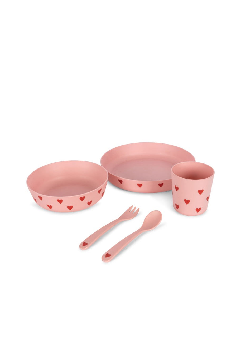 A pink dinner set for children including a plate, bowl, cup, and cutlery, all adorned with small red heart patterns.