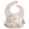 A silicone baby bib in pink with a floral pattern, featuring adjustable neck fasteners and a deep front pocket.